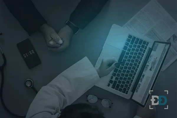 Seen from above, a clinician in a white coat points a pen at a laptop screen, with a stethoscope and a phone on the desk.