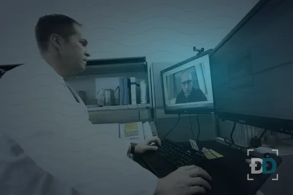 A doctor in a white coat types at his desk while a patient talks to him on a video call on the monitor.