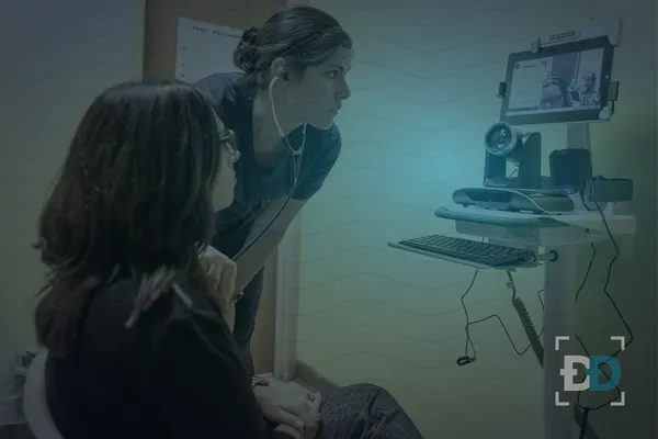 A nurse listens with a stethoscope as a seated patient waits beside a telehealth cart with a camera and screen.