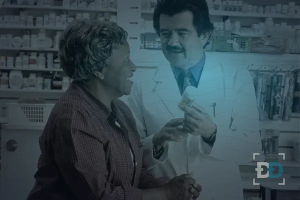 A pharmacist in a white coat holds up a pill bottle and explains a prescription to an older woman in front of stocked shelves.